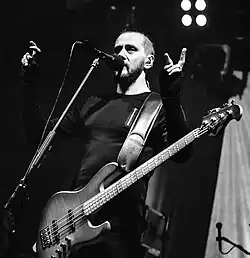 black-and-white image of Mariusz Duda standing onstage, appearing to speak or sing into a microphone, with both arms raised to shoulder height, electric bass guitar around his neck