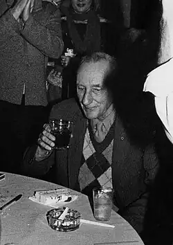 A black-and-white photograph of an older man sitting at a table and drinking a glass, surrounded by people