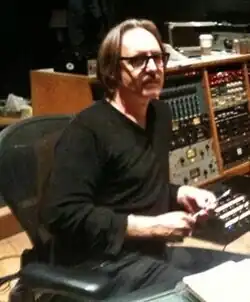 A man wearing glasses and black clothes sits in the control room of a recording studio.
