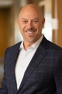Had and shoulders picture of a smiling, middle-aged white man with a clean-shaven head and trimmed beard with pepper-grey hair. He appears to have hazel eyes and is waiting a blue-plaid sports jacket over a white button-up shirt with the top button unbuttoned.