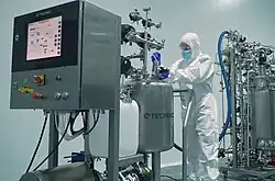 A person in a full white cleanroom suit, blue gloves, and a face mask operates complex stainless steel bioprocessing equipment in a sterile lab