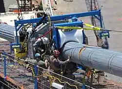 A blue machine is on a bridge being constructed; the machine has roller wheels that are pressing on a steel cable.
