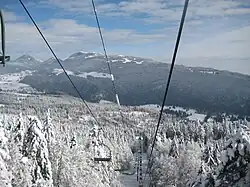 A winter mountain landscape in France with a chairlift ascending over snow-covered forests and distant peaks under a blue sky.