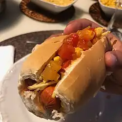 In Brazil, a cachorro quente is served on a bread roll with a tomato-based broth, corn, and potato sticks.