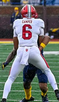 Camper wearing a white Indiana Hoosiers football jersey with the picture showing backside showing his name, Camper, and number, 6, in red text. He is against Michigan in 2023 and also is wearing a red helmet with a white stripe down the middle.