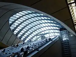 Escalators descending into an underground station with a glazed canopy above, with a glazed lift shaft on the side