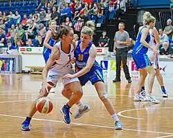 A female basketball player is attempting to drive to a basket while another female player is guarding her, and attempting to reach for the ball.