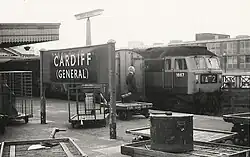 A black-and-white photograph of a railway platform. Two trains stand side by side on the image's right while a sign on the platform reads "Cardiff (General)". A formally dressed gentleman stands on the platform, looking away from the viewer. A briefcase rests on a small trolley beside him