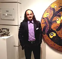 Man with long curly hair wearing a dark suit and lavender shirt standing beside a circular abstract artwork in a gallery