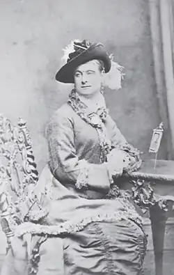 A woman in a dress and hat sitting in a chair, looking slightly away from the camera