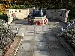 Slab paved walkway leading to wider paved area with a central compact raised plinth topped by three cones representing armed forces on land, sea and in the air, backed by a natural stone arced low wall having individual inscribed plaques