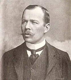 Head and shoulders portrait of a man with receding hair, heavy moustache, looking left from the image. He wears a high white collar, black necktie, dark waistcoat and jacket.