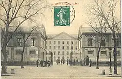 An entrance gate between two gatehouses, opening onto a vast courtyard, the latter enclosed in the background by a four-storey building.