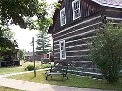 A small log building made of dark brown horizontal timbers with white-trimmed windows sits beside a paved walkway. A wooden bench and green shrubs are nearby.