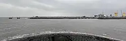 A modern quayside at a harbour entrance on a grey day, to the right large parts for offshore wind foundations are stacked behind the harbour building. At the bottom, the stone blocks of the old breakwater are visible, with waves breaking on them