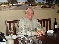 A man with white hair and glasses wearing a brown button-down shirt, smiling at the camera