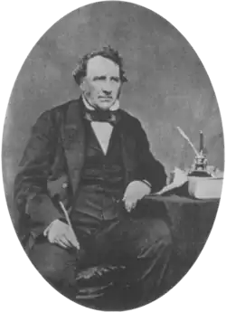 A middle aged Victorian gentleman sits beside a table wearing a dark suit with waistcoat and bow tie. His left elbow rests on the table. In his right hand he holds a quill pen.