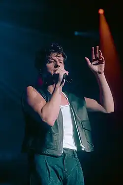 A white male in a tank top and sleeveless jean jacket speaks into a microphone.