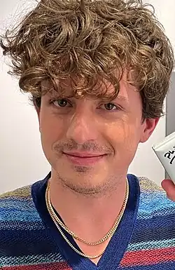 Puth with a full front of curly brown hair, closed-mouth smile, and stubble has two chain necklaces and a colored horizontally striped shirt.