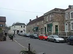Street scene showing road junction and grey stone buildings with parked cars in front of them. To the left is a grassy area with a tree.