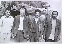 Black-and-white photograph from the 1970s showing four men standing outdoors in Bigherd, Iran; the village’s last chief stands second from the left, with palm trees and a vehicle visible in the background.
