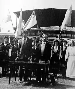 Chornovil and another individual standing in front of several women dressed in Ukrainian traditional clothes, and below multiple Ukrainian flags