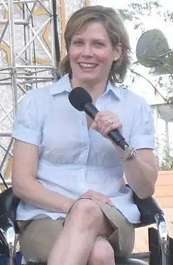 Smiling woman in a light blouse sits outdoors with a microphone in hand.