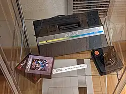 Closeup of a black flat electronic device with a receptacle slot flanked on both sides by sets of two switches. In front of the device is a stack of two black plastic video game cartridges. The top one's label reads "Atari 26000 Video Game Cartridge: Joust"