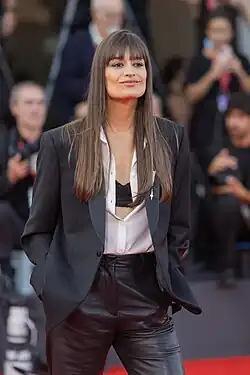 Clara Luciani wearing a white, unbuttoned shirt partly revealing black brassiere, with black jacket and black leather pants; she is standing on a red carpet, surrounded by blurry photographers, and looking left and slightly up of camera with a pouty smile, hands in jacket pockets.