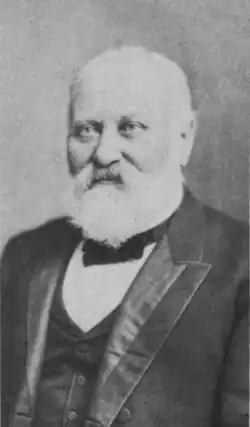 portrait shot of a man in late middle age with white hair and beard. He is wearing a suit with vest and a small bow-style tie.