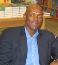 A man, wearing white shirt with a logo and the word "NBA PLAYERS" on the front, is standing and posing for a photo.