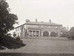 Black-and-white photograph of a mansion in parkland, with a portico on the left-hand side of the façade