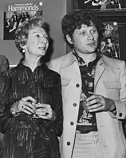 A black-and-white 1976 image of actress Jean Anderson on the left and Colin Baker on the right, both holding a drink