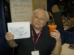Colin Baker at an autograph signing in 2009 looking at the camera; in his left hand is a notepad with "Doctor Who Forum" written on it; in his right hand is a cup filled with carrot juice.