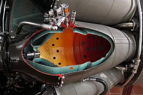 Rolls-Royce Nene with nine combustion chambers. The cutaway is one of 2 chambers fitted with a flame igniter which places the igniter in a cooler location than directly in the hot gas stream. During a start atomized fuel from the small self-contained unit (orange-coloured solenoid shown) is ignited by its ignition plug and the flaming jet of fuel is projected into the main fuel spray from the burner. Combustion is propagated to all the chambers through interconnecting tubes.[88]