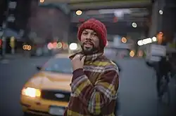 A man with facial hair, dressed in a flannel jacket and knitted hat, and a city street in the blurred background