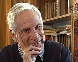 Colour photograph of a clean-shaven, smiling, white-haired man, resting his chin on his hand, with book shelves in the background