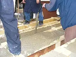 A handheld concrete vibrator consolidates fresh concrete in wooden formwork for a concrete beam.