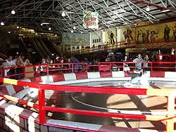 Interior of a large funhouse. In the foreground are riders sitting on a spinning wooden disc.