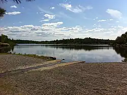 Entrance to Lake Laurentian, showing a rocky beach, lake and boat ramp surrounded by trees under a blue sky.