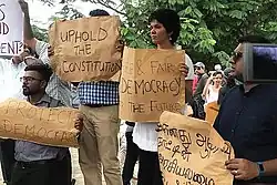 Shreen Abdul Saroor stands in the centre of a crowd at a protest. She holds a cardboard sign that reads "Free & Fair Democracy For the Future". Men on her left hold other cardboard and paper signs reading "Uphold the constitution" and "Protect democracy". A man to her right holds a sign written in Sinhala.