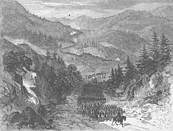 A lithograph in black and white of a road winding through a mountainous region. An army of thousands is marching along the road, led by a military officer on a horse.