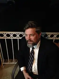 Schindler in a black suit and tie looking to his right while sitting on a chair on a patio or porch.