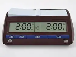 A brown chess clock with blue buttons along the bottom. A digital display shows the time remaining for each side