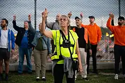 Margaret Dragu is in the foreground looking at her right hand and index finger which are held above her head. She has grey hair and is wearing high visibility equipment with a small camera strapped to her chest. Behind her a crowd of participants are facing in the same direction and holding the same pose. There is a chain link fence behind them.