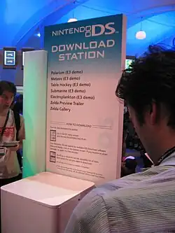 A kiosk with a large sign that fades vertically downward from light blue to white. The words "Nintendo DS Download Station" are above a list of demo names, which includes "Polarium (E3 demo)".