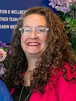 Headshot of Marlowe smiling in front of a floral background, wearing a red blazer, black shirt, and gold necklace.