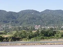 A highway with a metal guardrail runs across the foreground, with green trees and shrubs behind it. In the distance, a small town with scattered houses sits at the base of forested hills under a clear sky.