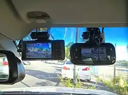 A pair of black devices with small monitors stuck with suckers to the inside of a car windshield with blue tint at the top, seen from inside the vehicle
