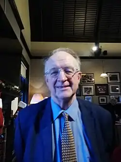 Clean-shaven White male with thinning white hair. He wears a blue suit, collar and tie, and glasses.